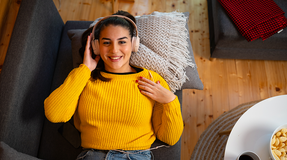 Birds eye view of a woman lying back on her sofa with headphones on listening to something and smiling